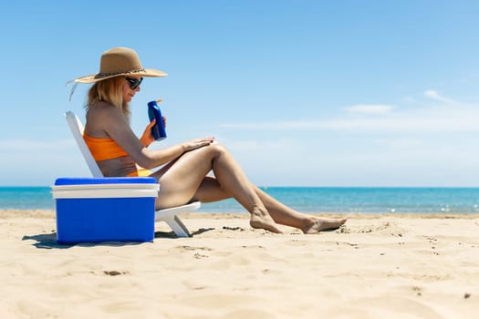 beach background with a girl, a slender girl in a swimsuit sits on a sun lounger smears her legs with sunblock, a blue portable refrigerator for drinks stands nearby. High quality photo