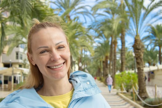 a girl of European appearance with blond hair, in a blue jacket, stands on the embankment, smiles around green palm trees. A beautiful seascape. High quality photo