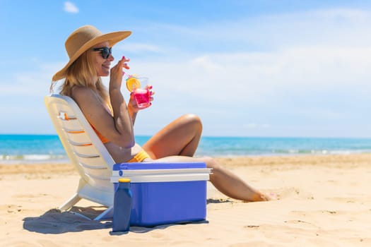 Happy young european girl with a cocktail in her hands is resting in a sun lounger on the sea near the blue refrigerator, there is space for an inscription. High quality photo