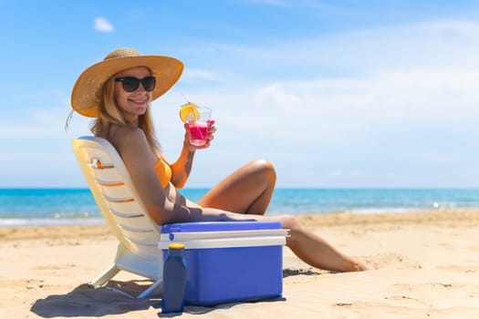 tanned girl in a swimsuit, hat and glasses sits on a sun lounger with a cocktail in her hands side view on the beach and there is a blue portable refrigerator for drinks next to it. High quality photo