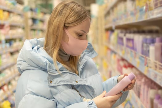 A girl with blond hair of European appearance in a blue jacket and a pink mask from coronavirus in a supermarket chooses shampoo