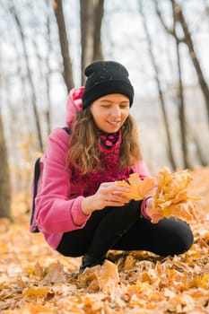 Portrait of beautiful happy young woman in pink jacket holding bouquet of yellow leaves at autumn park. Pretty Caucasian lady smiling and looking at camera during her walk outdoors. Generation Z and gen z youth.