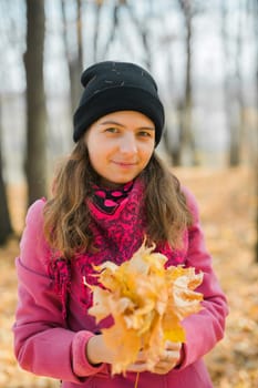 Portrait of beautiful happy young woman in pink jacket holding bouquet of yellow leaves at autumn park. Pretty Caucasian lady smiling and looking at camera during her walk outdoors. Generation Z and gen z youth.