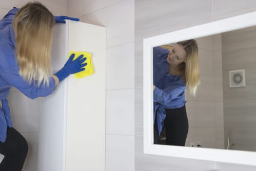 A young blond girl of European appearance wipes dust from white furniture with a yellow rag in the bathroom, wearing blue gloves. The girl can be seen in a white mirror.cleaning concept