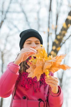 Portrait of beautiful happy young woman in pink jacket holding bouquet of yellow leaves at autumn park. Pretty Caucasian lady smiling and looking at camera during her walk outdoors. Generation Z and gen z youth.