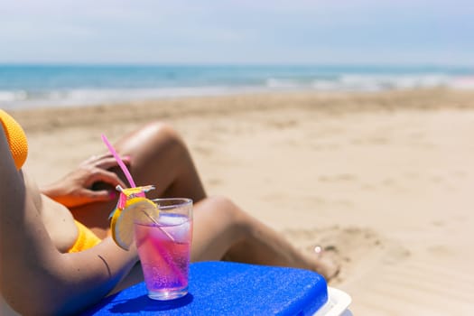 A tanned thin girl in a bikini sits on a sun lounger close-up on slender legs, there is a portable refrigerator, there is a glass with a cocktail on it, a beautiful beach landscape,High quality photo