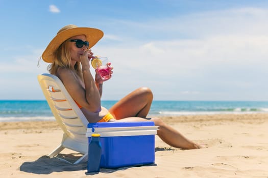 Happy young european girl drinks a cocktail and rests in a sun lounger on the beach, space for an inscription portable blue refrigerator by the sea. High quality photo