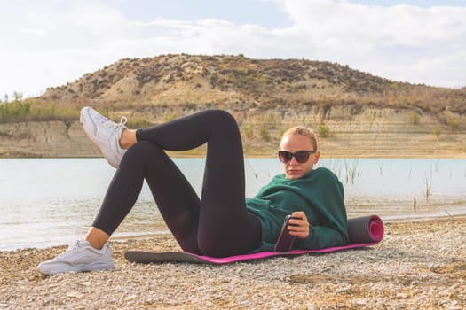 a girl of European appearance in sportswear lies on a yoga mat near a beautiful lake. Beautiful landscape of nature, there is a place for an inscription. Lake with turquoise water. High quality photo