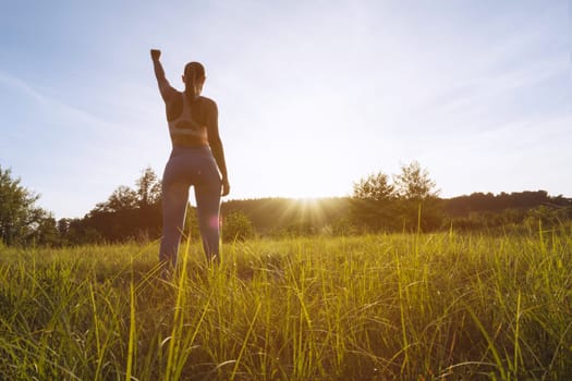 girl in a fitness suit stands with her back to the camera at sunset with her hand raised up after a workout in the fresh air, motivational girl silhouette place for an inscription. High quality photo