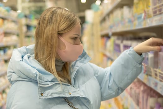 A girl with blond hair of European appearance in a blue jacket and a pink mask against viruses in a supermarket chooses household goods. High quality photo