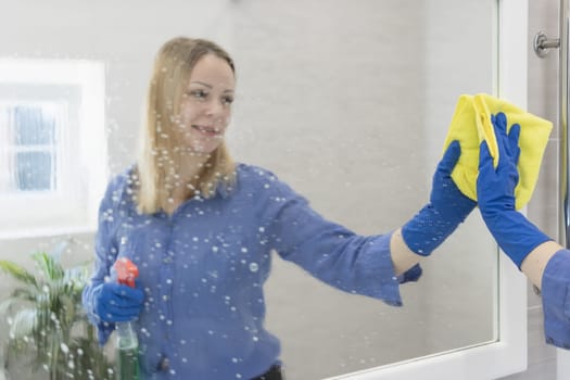 happy blond girl with European appearance washing mirror with yellow cloth in blue gloves at home.House cleaning concept