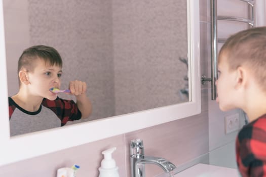 A small blond boy of European appearance learns to brush his teeth in a home bath in red pajamas and looks into a white mirror. The child learns to stay healthy. healthcare concept.