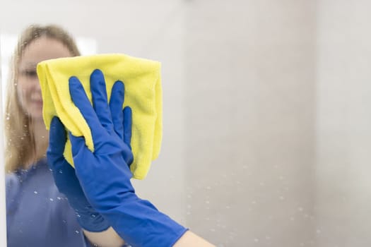 happy blond girl with European appearance washing mirror with yellow cloth in blue gloves at home.House cleaning concept