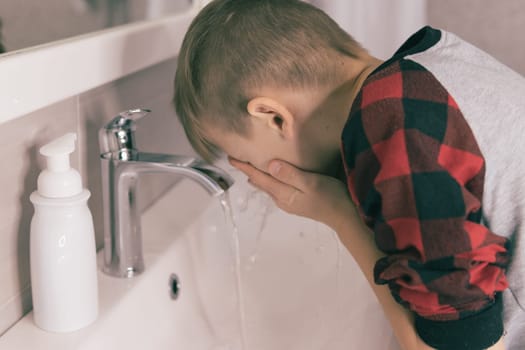 a small blond boy of European appearance washes himself in the bathroom in a white wash basin, in pajamas. There is white soap on the sink. The concept of a healthy lifestyle