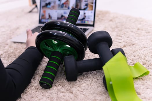 Fitness accessories at home on a carpet close-up, dumbbells, an elastic band for sports, a press roller. In the background, a girl is watching home workouts in a laptop. the concept of sports