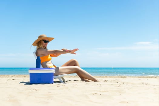 a tanned girl in a swimsuit, hat and glasses sits on a sun lounger smears sunscreen on her hands side view on the beach and stands next to a blue portable refrigerator for drinks. High quality photo