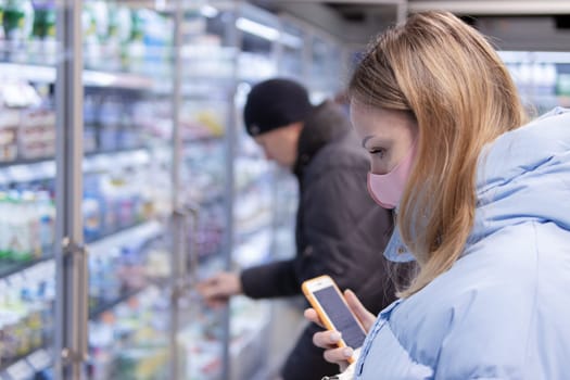 A girl with blond hair in a blue jacket holds a phone in her hands, wears a pink medical mask from coronavirus while shopping in a supermarket - health, safety and pandemic concept - a girl in a protective mask stocks up on food.