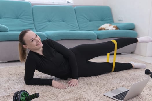 a young smiling girl of 30-40 years old, of European appearance with blond hair tied in a ponytail in a black sports uniform, goes in for sports with a yellow sports elastic band at home, lying on the floor on a white carpet with a laptop and smiling, a roller for the press and dumbbells lies on the floor nearby. In the background lies a white cat on the couch.Home workout concept