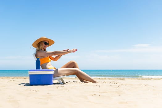 a tanned girl in a swimsuit, hat and glasses sits on a sun lounger smears sunscreen on her hands side view on the beach and stands next to a blue portable refrigerator for drinks. High quality photo