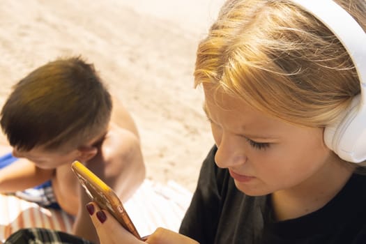 teenagers boy and girl are sitting on the beach, the girl is wearing headphones and with a phone in her hands listening to music, the boy is lying sunbathing. High quality photo