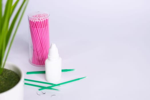 Materials for eyelash extension lie side by side on a white background, pink and green microbrushes, in a white jar of liquid for eyelash extension. On the right there is a place for an inscription.