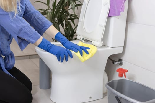 a girl of European appearance with fair hair in blue gloves is cleaning the bathroom in the house and cleans the toilet bowl with a yellow napkin