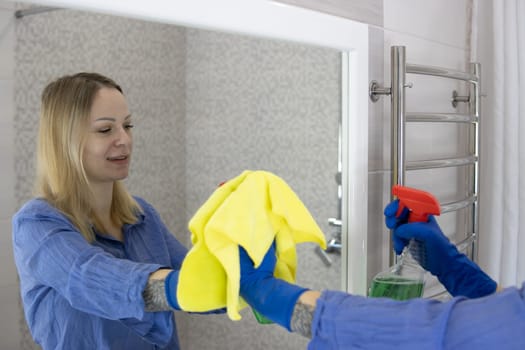 happy blond girl with European appearance washing mirror with yellow cloth in blue gloves at home.House cleaning concept