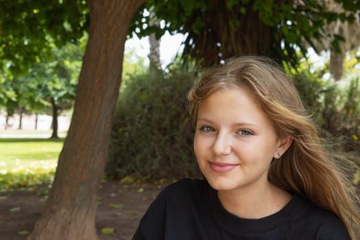 portrait of a teenage girl with blond hair, the girl sits in the park and looks at the camera. High quality photo