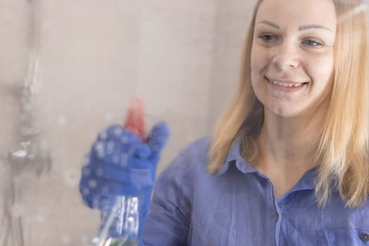 happy blond girl with European appearance washing mirror with cleaning spray in blue gloves at home.House cleaning concept