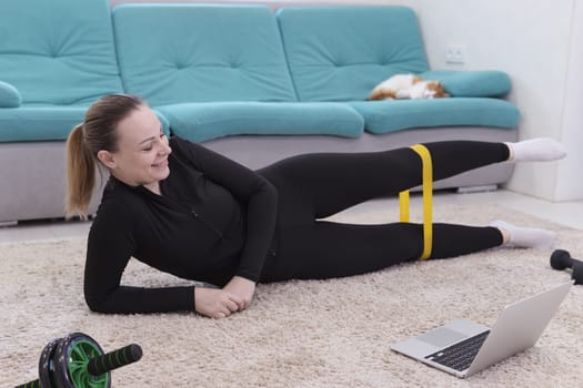 young smiling girl of European appearance with blond hair in a black sport uniform goes in for sport with a sport elastic band at home lying on the floor in a laptop.The concept of home training