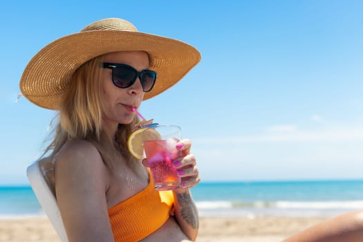 Portrait of a happy european woman in an orange swimsuit and a hat and glasses sunbathing on a plastic sun lounger on the beach.Girl with her hand raised up is sunbathing on the sea.High quality photo