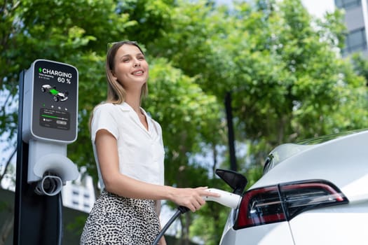 Young woman travel with EV electric car charging in green sustainable city outdoor garden in summer. Urban sustainability lifestyle by green clean rechargeable energy of electric BEV vehicle innards