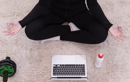 Sporty healthy calm woman sitting on mat in lotus position,wearing sporty black uniform,watching online yoga class,meditate,do breathing exercises on laptop.White bottle with water is standing nearby