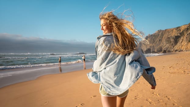 Woman running on beach smiling for camera, wearing sweatshirt and beach shorts. Woman walks along beach, smiling enjoying vacation against blue sky and mountain on background