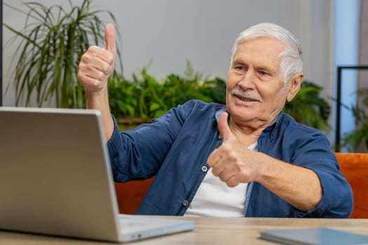 Senior man making video webcam conference call with friends or family, shows thumbs up, enjoying pleasant conversation at home apartment indoors. Elderly mature grandfather sits on couch at table desk