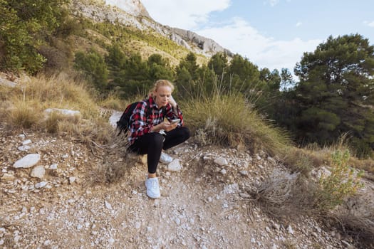 a girl in sportswear sits on a stone, rests from a hike in the mountains, a girl sits looking at the phone. High quality photo