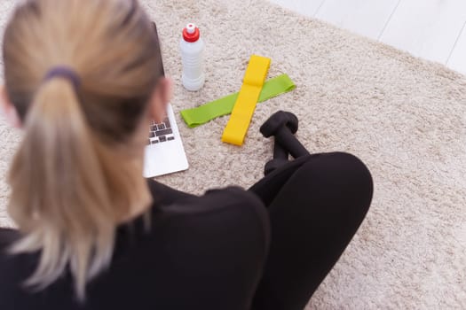 A sports girl sits and rests after training at home online on the floor nearby lies a close-up of a laptop, water in a white bottle, colored rubber bands for squats and black dumbbells