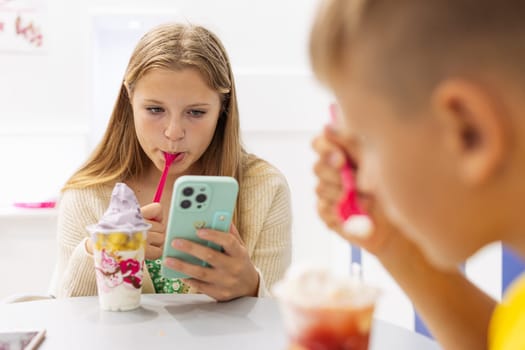 brother and sister are sitting in a cafe, eating ice cream and a girl is looking at a message on the phone, the concept of a summer family vacation. High quality photo