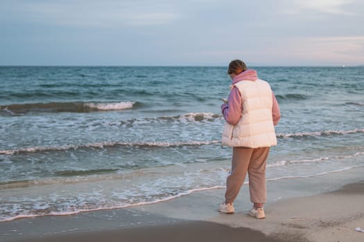 A girl of European make in casual clothes stands near the ocean with a phone in her hands is sad and looks into the phone. High quality photo