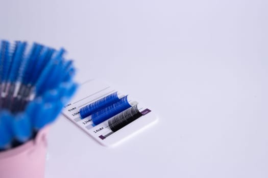 Artificial eyelashes in black and blue are glued on a white tablet on a white background. There is a for an inscription on the right.Blurred blue brushes in front of the eyelashes