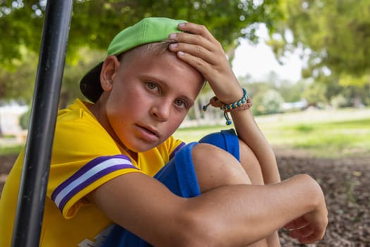 a boy of European appearance in a yellow T-shirt and a green cap sits in the park with a red face and a tired look, the child plays active games in the fresh air. High quality photo