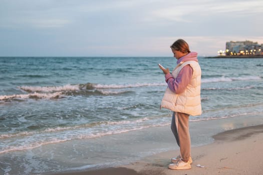 Portrait of an excited girl in her hands with a phone, standing on a beach with a blue sea. Sad and depressed young woman standing alone on the beach by the sea. Depression concept. High quality photo