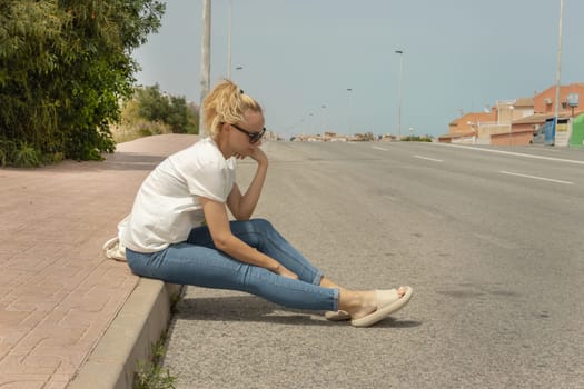 a sad girl sits on the sidewalk in jeans and a T-shirt and glasses, looks at the road, there is a place for an inscription. High quality photo