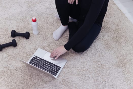 Athletic middle-aged woman in black sportswear sits on the floor on a carpet with dumbbells and a bottle of water and uses a laptop at home in the room. The concept of sports and recreation.