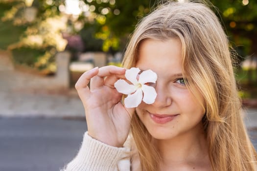 portrait of a teenage girl of European appearance, blonde hair, the girl stands on the street holding a white flower near her eye there is a place for an inscription. High quality photo