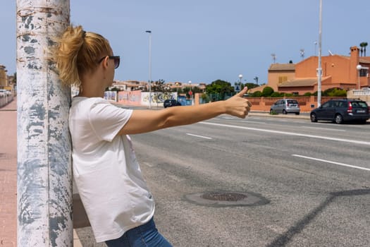 a beautiful girl in a white t-shirt and jeans stands on the road with her back to the camera and stops the car on the road, there is a place for an inscription. High quality photo