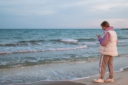 A girl of European make in casual clothes stands near the ocean with a phone in her hands is sad and looks into the phone. High quality photo