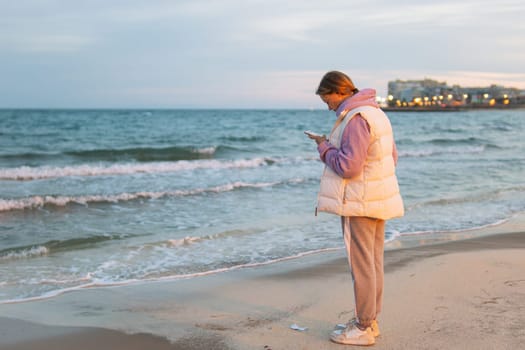 A girl of European make in casual clothes stands near the ocean with a phone in her hands is sad and looks into the phone. High quality photo