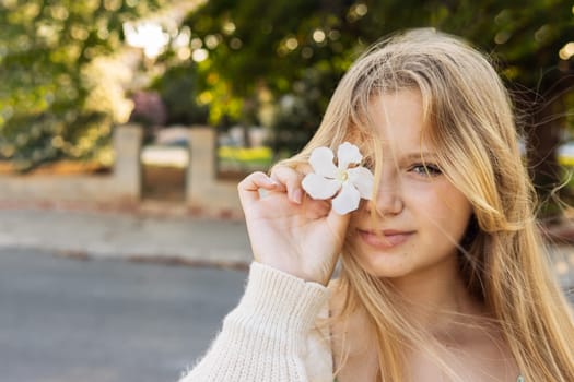portrait of a teenage girl of European appearance, blonde hair, the girl stands on the street holding a white flower near her eye there is a place for an inscription. High quality photo