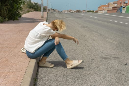 a girl sits on the pavement in jeans and a T-shirt, turned her back to the camera, looks at the road where cars drive. High quality photo